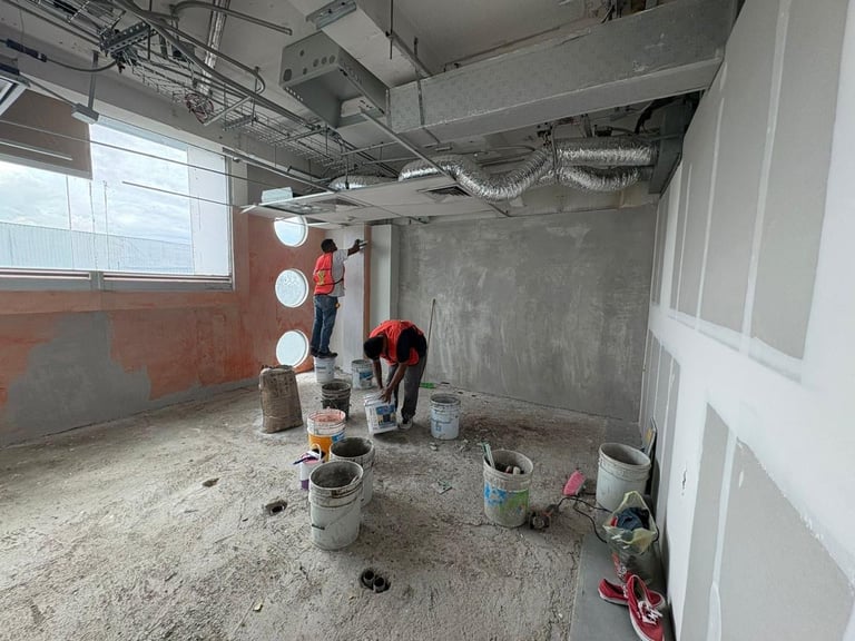 Workers in construction area with concrete floor, temporary lighting, buckets, and tools under exposed ceiling ductwork