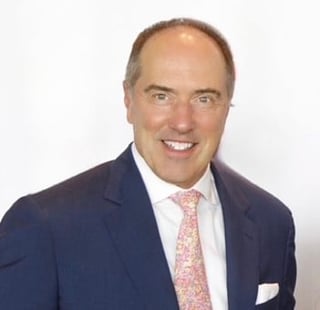 Professional headshot of a middle-aged man in a navy blazer, white shirt, and pink tie, smiling against a white background.
