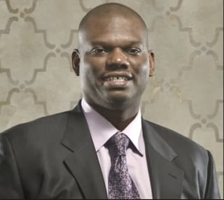 Professional headshot of a man in a dark suit and lavender shirt with purple tie, smiling at the camera against a patterned gray background.