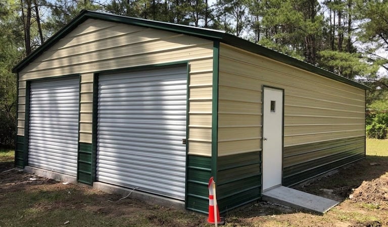 Metal garage with tan and green siding, two white roll-up doors, single entry door, surrounded by trees