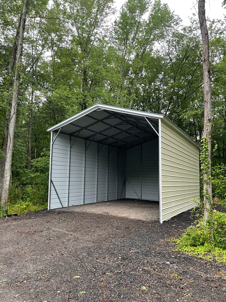 Metal carport with white and gray exterior surrounded by tall trees on gravel ground
