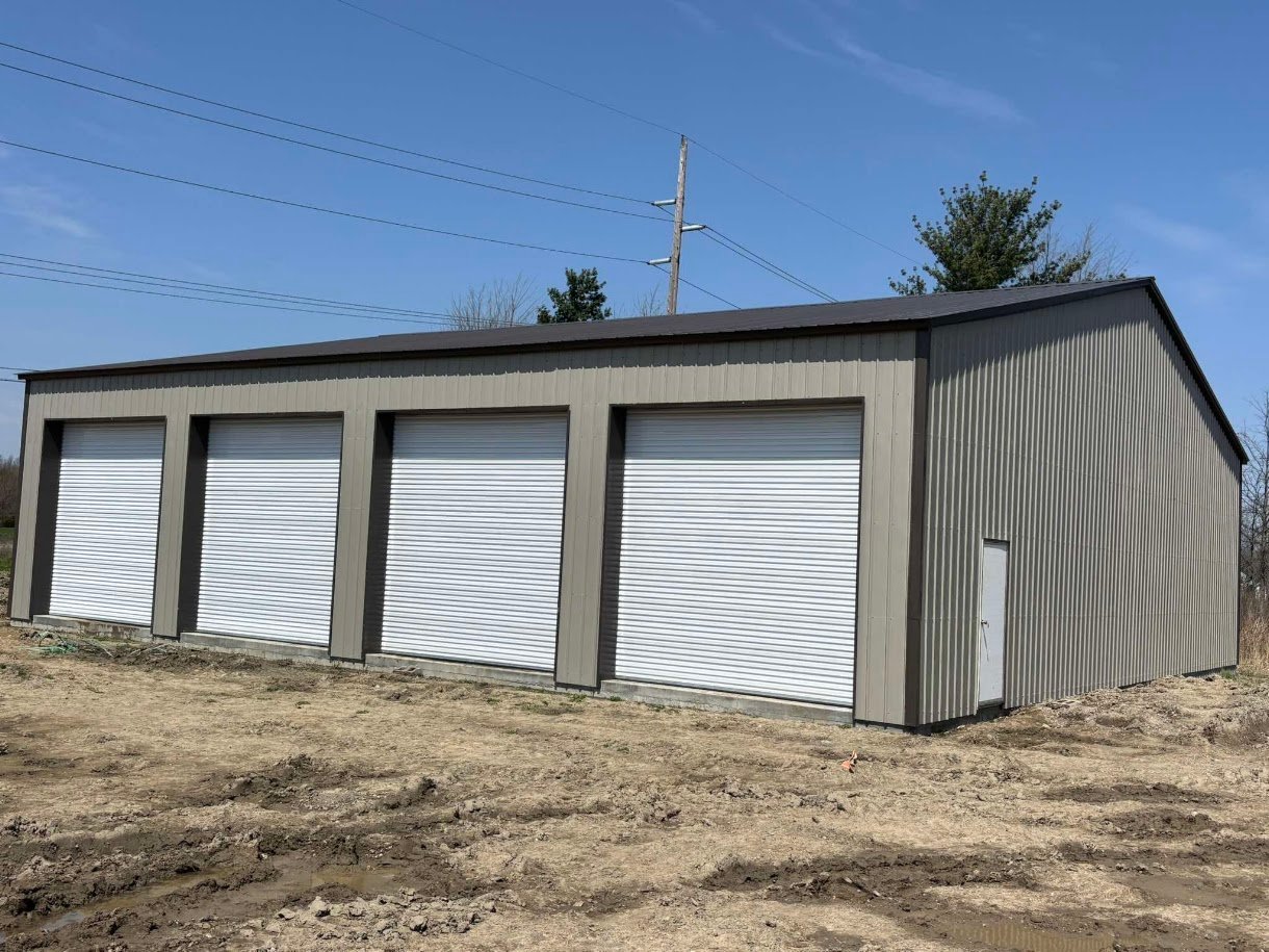 Modern metal garage building with four white roll-up doors, corrugated steel exterior, on dirt lot with utility pole and bare trees in background
