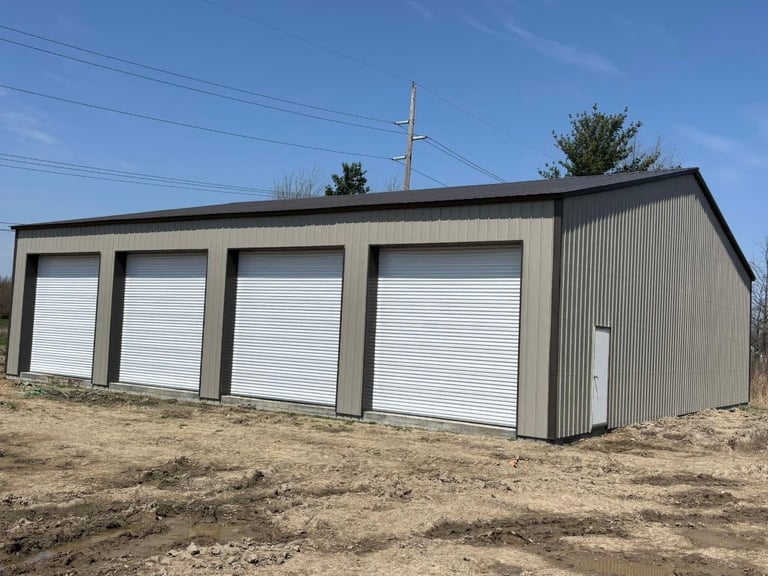 Modern metal garage building with four white roll-up doors, corrugated steel exterior, on dirt lot with utility pole and bare trees in background