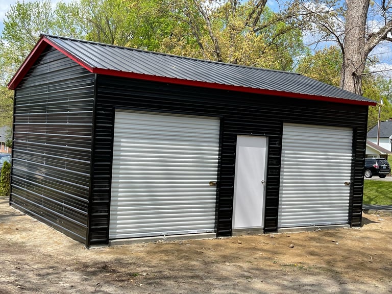 Black metal storage building with red roof, white garage doors, and white entry door surrounded by trees