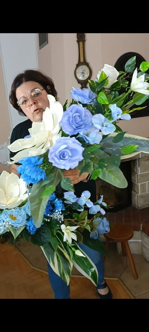 Osoba w okularach trzymająca duży bukiet białych, niebieskich i fioletowych kwiatów z zielenią w domu