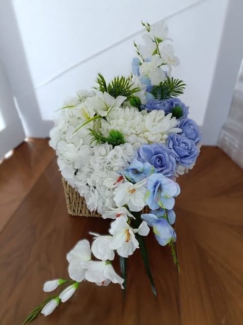 Elegancka kompozycja kwiatowa w wiklinowym koszu z białymi różami, niebieskimi różami, białymi orchideami i zielenią na drewnianej powierzchni