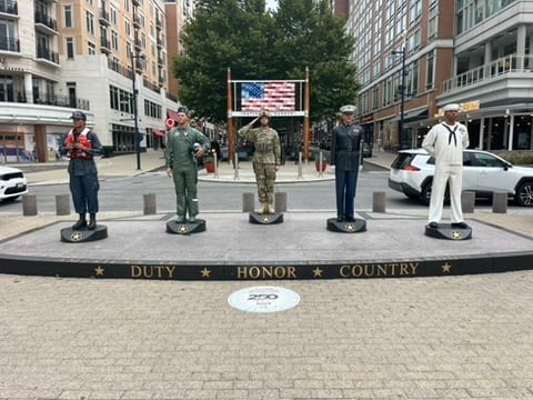 Five military and civilian figurines stand on a street display with Duty, Honor, Country inscribed on the base, with city buildings in background