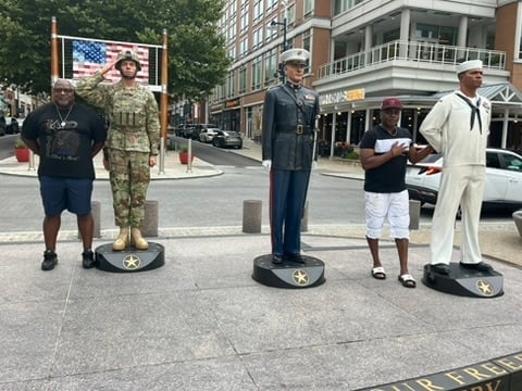 Five life-sized cardboard cutout figures standing on a city street, depicting military and civilian personnel in various uniforms