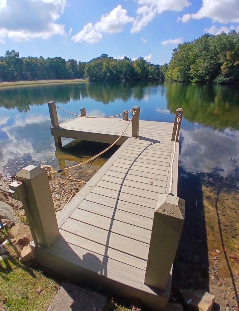 Wooden dock extending into calm lake with moored boat, surrounded by dense green trees and blue sky with white clouds