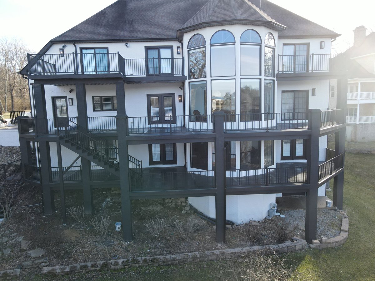 Large elevated waterfront house with white and dark exterior, multiple decks, and expansive windows, built on stilts over landscaped grounds