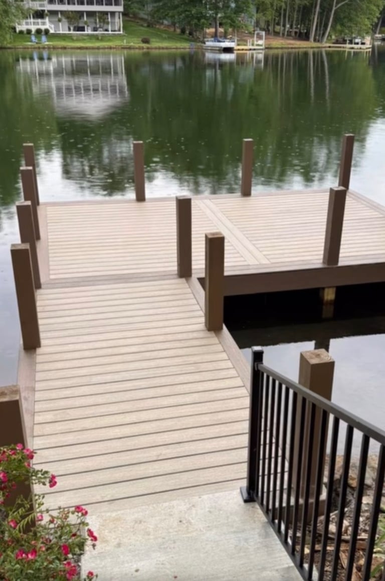 A wooden dock with pilings extends into calm green water, with waterfront homes visible across the lake and flowering plants in the foreground