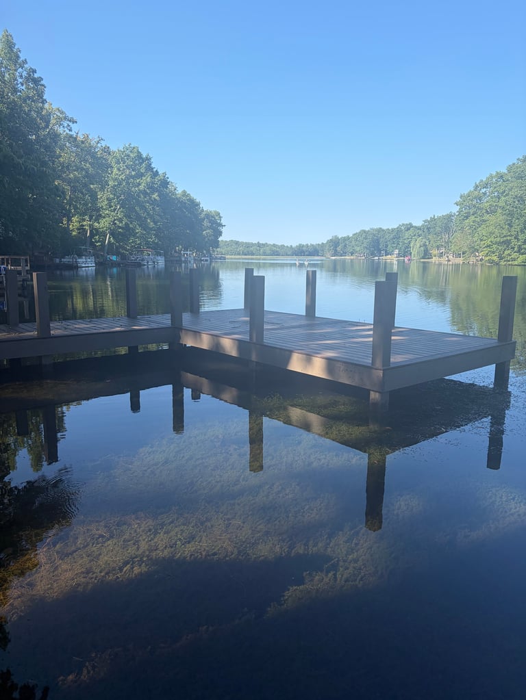 Wooden dock extending into calm, mirror-like river surrounded by dense green trees under clear blue sky