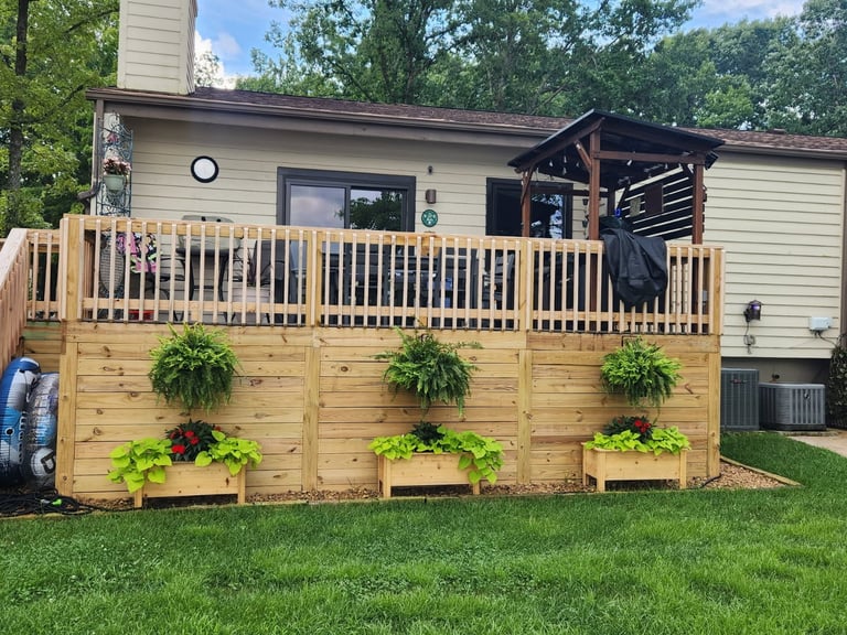 A wooden deck with white railings attached to a beige house, featuring three hanging planters with green foliage along the deck base