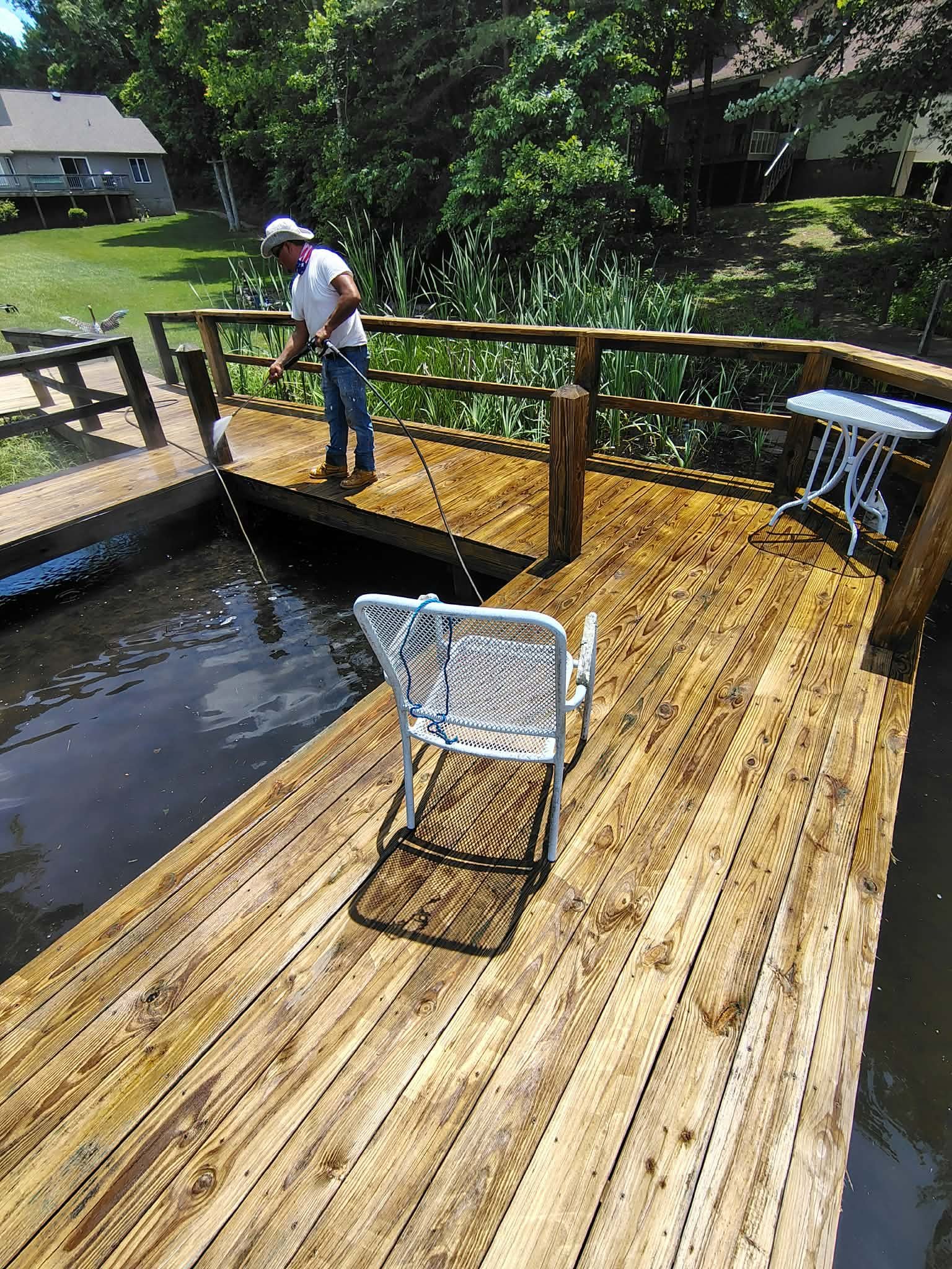 Person fishing on a wooden dock extending over a pond, with a chair in foreground and residential buildings visible in background