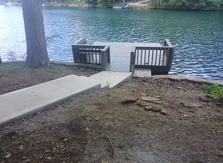 Concrete dock with railings extending into green water, with trees and waterfront homes visible across the lake