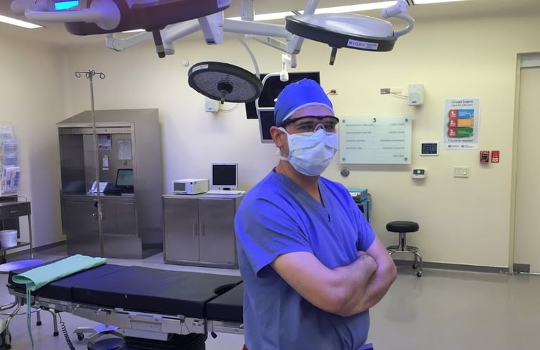 Surgeon in blue scrubs and surgical mask standing in operating room with overhead surgical lights and medical equipment