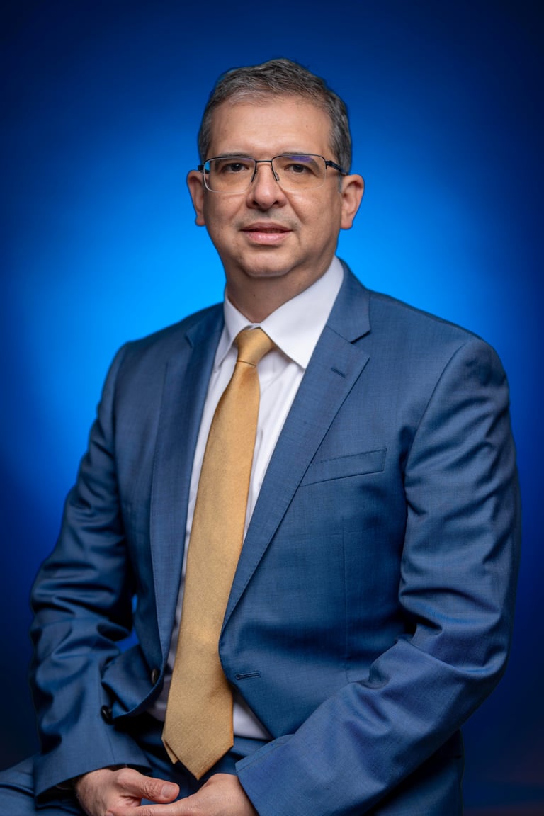Professional headshot of a middle-aged man wearing a blue blazer, white dress shirt, and gold tie against a blue background.