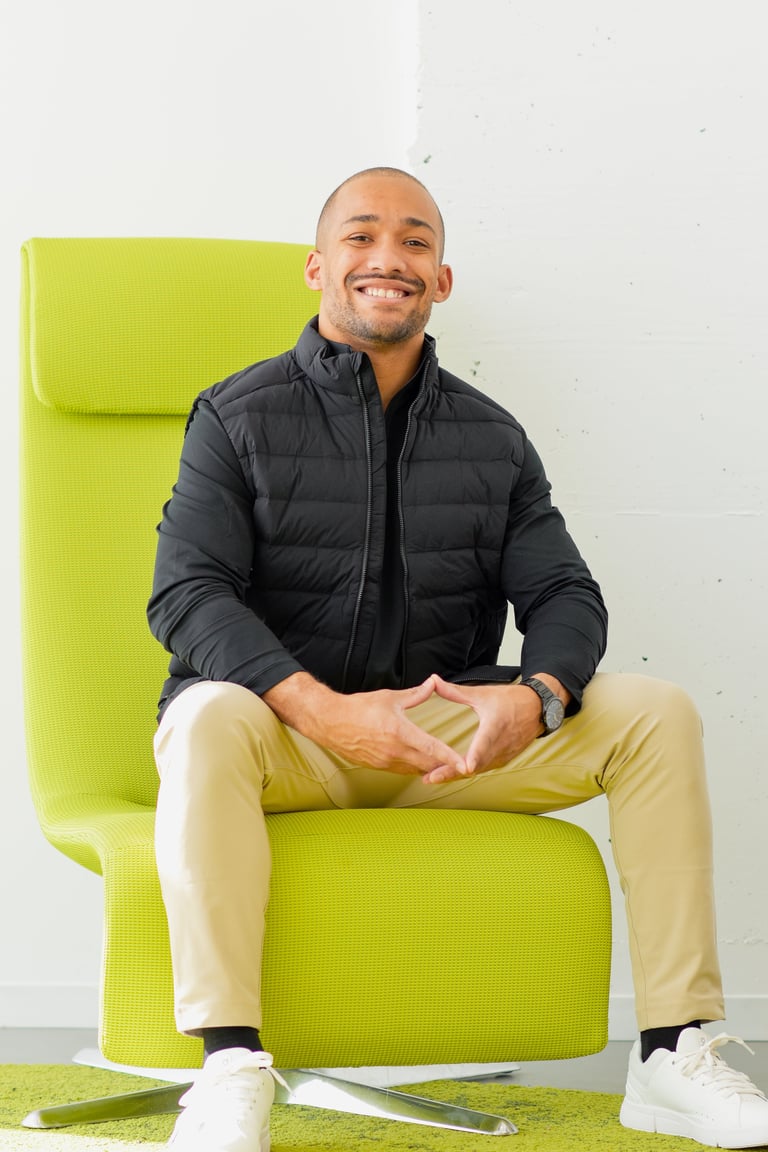 Smiling man sitting on a bright yellow chair wearing a black puffer jacket and tan pants with white sneakers