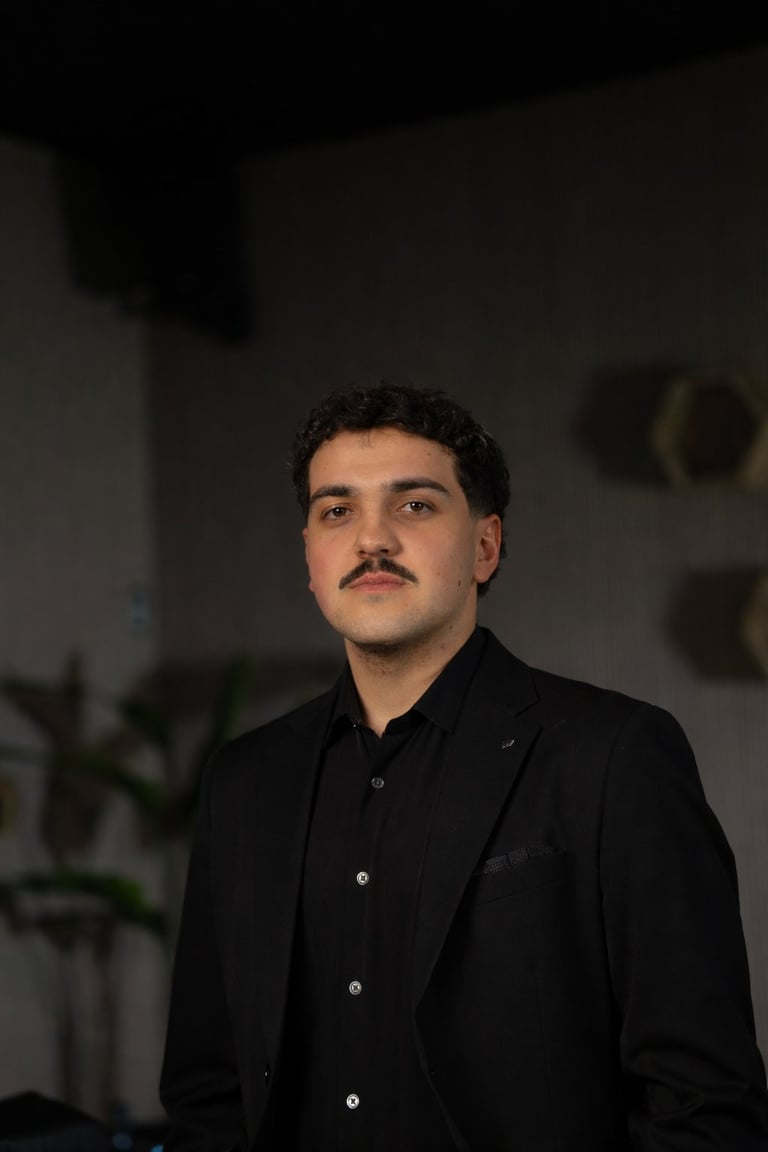 William Couturier — Man with dark hair and mustache wearing black shirt and blazer in dimly lit interior setting