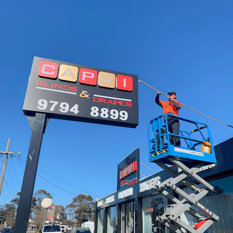 Worker on hydraulic lift maintaining colorful business signage advertising blinds and drapes against clear blue sky