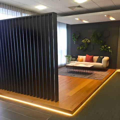 Modern office lounge space with black vertical blinds, beige sofa, colorful pillows, potted plants, and warm LED floor lighting