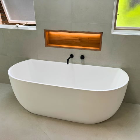 Modern white freestanding bathtub with black faucet and wooden niche shelf in minimalist bathroom