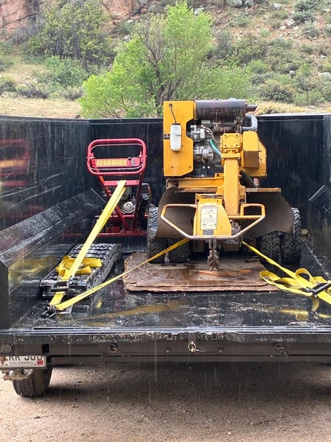 Yellow Vermeer stump grinder and red tracked equipment secured with yellow straps on a flatbed trailer in a Colorado canyon setting