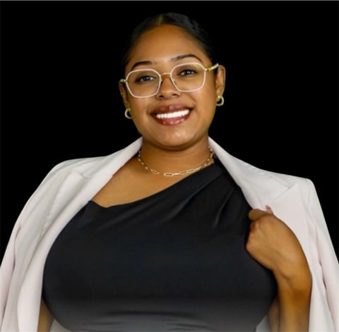Woman wearing glasses and a white blazer over a black top, smiling at the camera against a black background