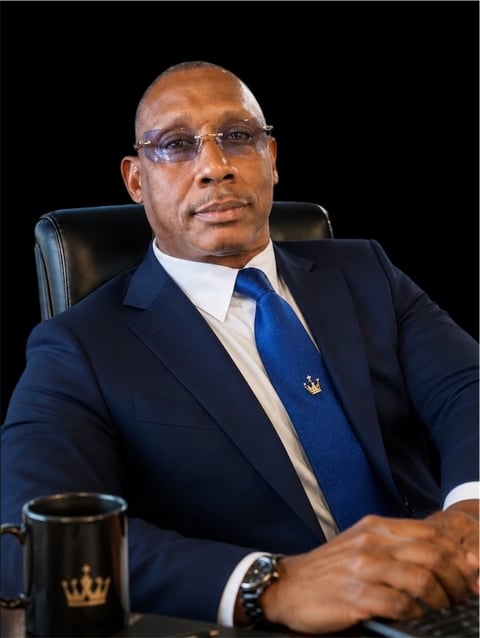 Professional portrait of a man in dark business suit and blue tie seated in office chair against black background
