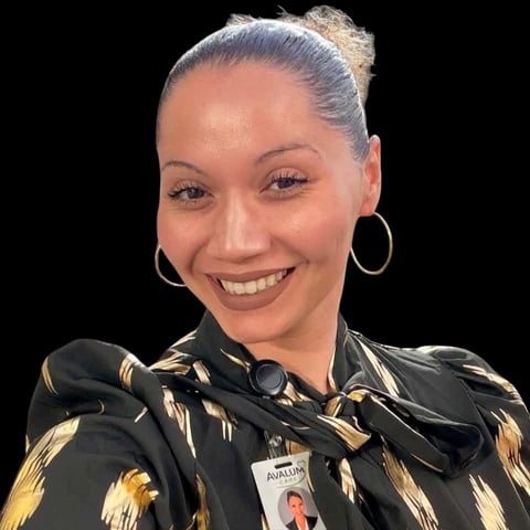 Smiling woman with blonde hair in a bun wearing gold hoop earrings and a black and gold patterned jacket against a black background