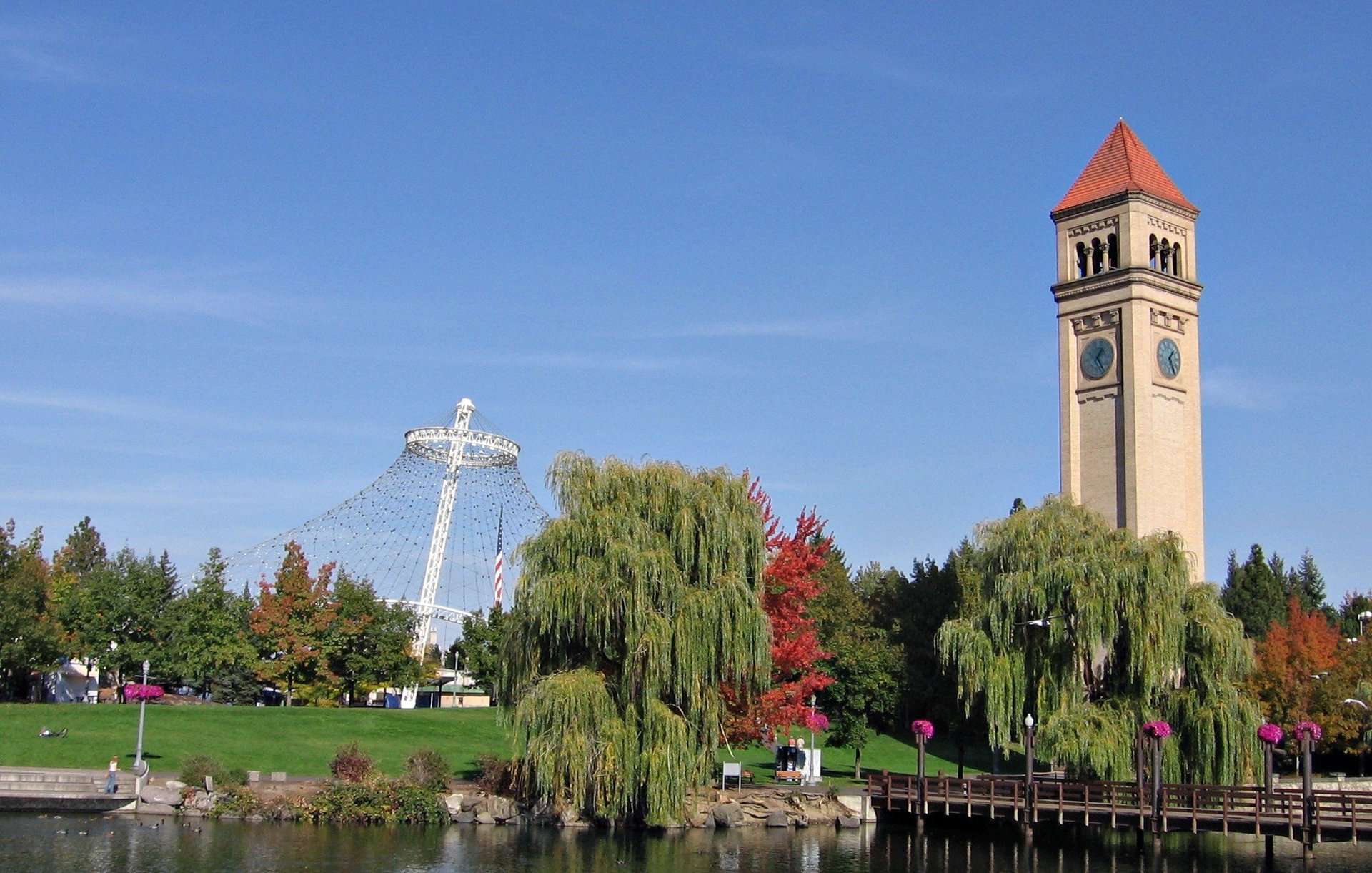 Riverfront cityscape with white cross monument, clock tower with orange roof, and weeping willow trees along grassy park and water