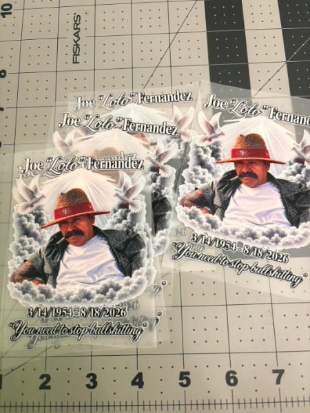 Memorial stickers featuring Joe Lolo Fernandez with red hat and clouds, dated May 1954-August 2026, on pegboard with ruler