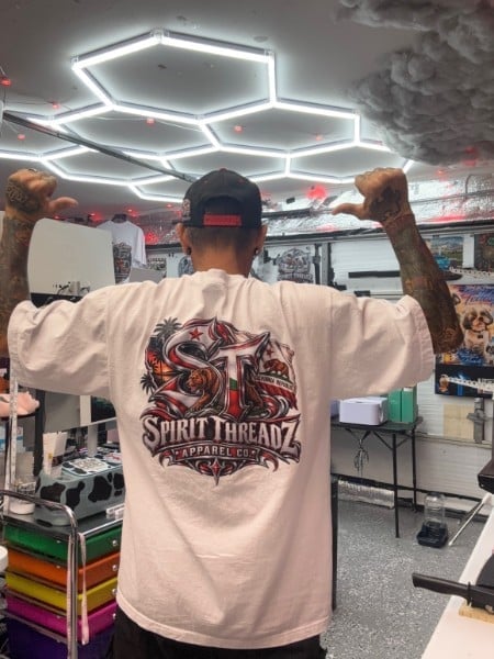 Person from behind wearing Spirit Threadz white t-shirt with arms raised in a tattoo shop with neon lighting
