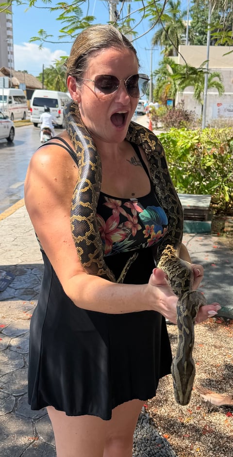 Woman wearing sunglasses holding a python snake around her shoulders outdoors, smiling with mouth open
