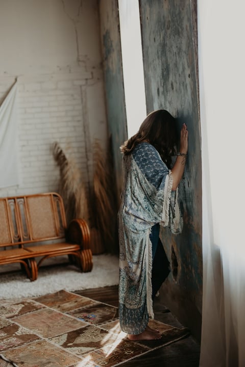 Woman leaning against weathered gray wall in ornate shawl, wooden chair and patterned floor visible behind