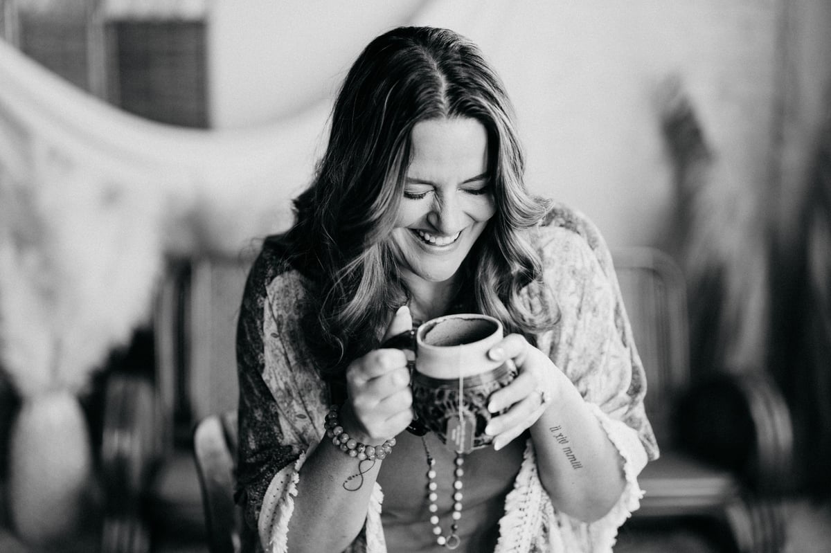 Black and white portrait of a smiling woman holding a cup, wearing a patterned cardigan and necklace