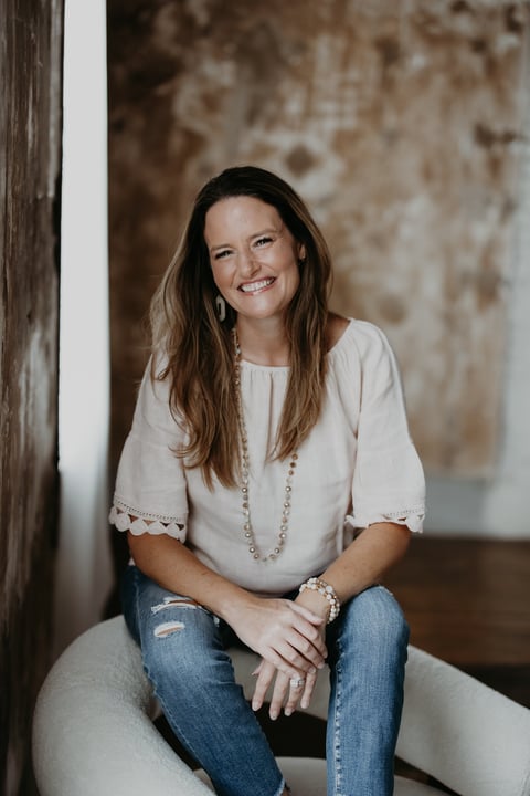 Woman with long brown hair wearing a cream top and jeans, sitting against a rustic textured wall, smiling at the camera