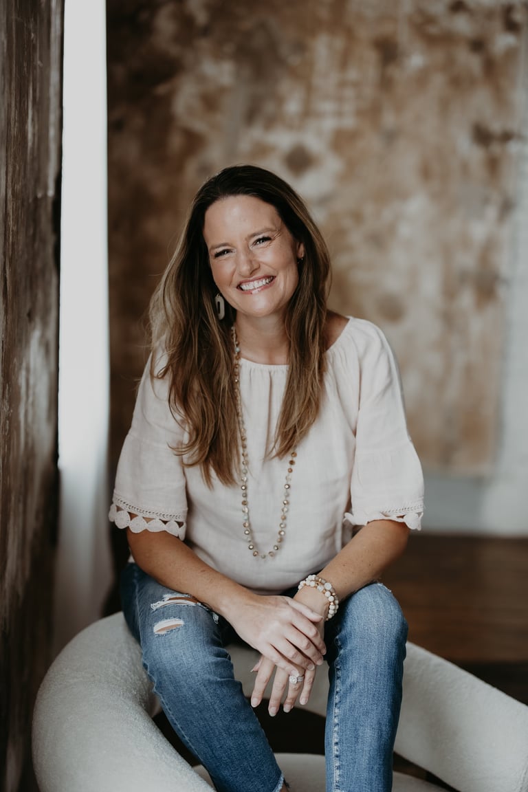 Woman with long brown hair wearing a cream top and jeans, sitting against a rustic textured wall, smiling at the camera