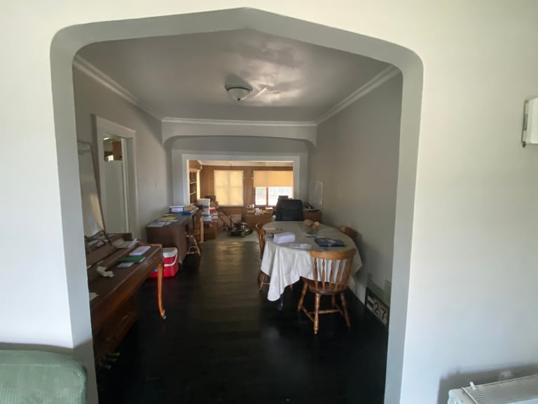 Interior view of a home showing a hallway leading to a living room with furniture, wooden dresser, dining table, and window with warm natural light