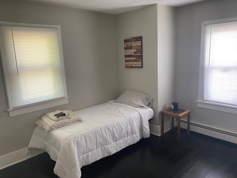 Minimalist bedroom with a twin bed, white bedding, wooden nightstand, and two windows with blinds on a dark floor