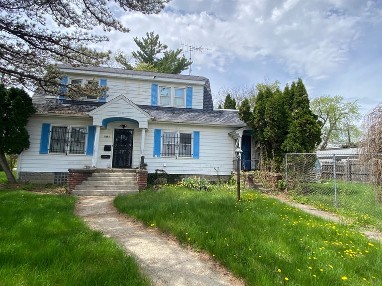 White house with blue shutters and trim, brick chimney, front lawn with yellow wildflowers, metal fence, and mature trees
