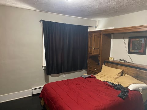 Bedroom with queen bed covered in red comforter, black curtains on window, wooden headboard with shelving, and dark flooring