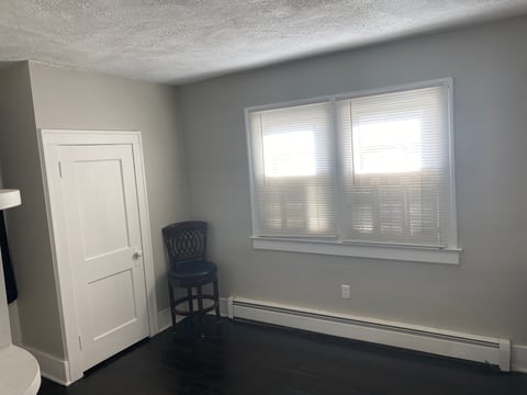 Empty room with gray walls, white door on left, two windows with blinds, dark floor, and single blue chair
