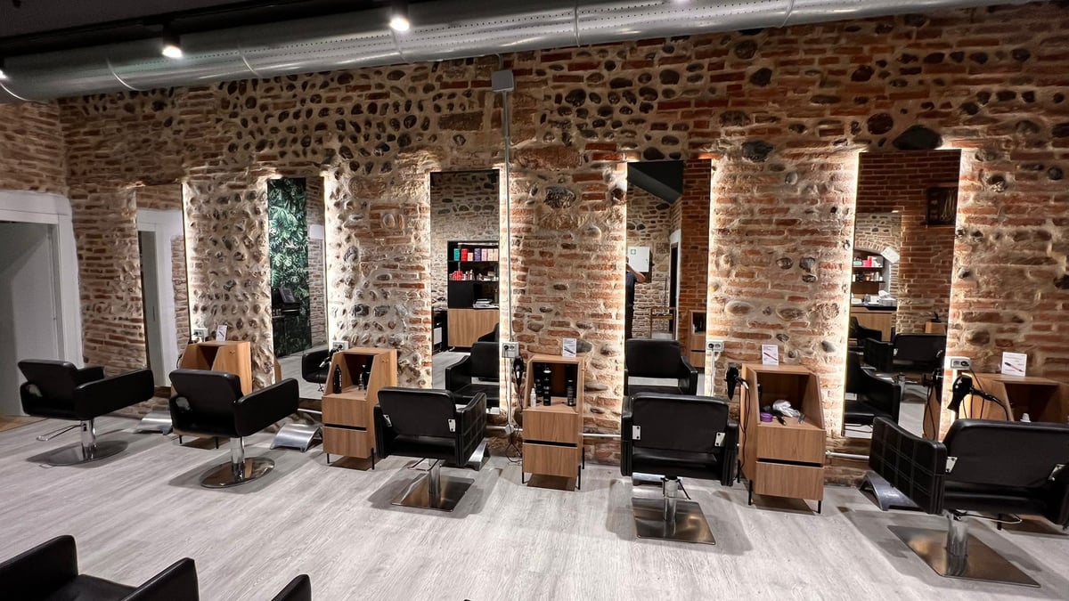 Modern hair salon interior with exposed brick walls, multiple styling stations with black chairs, and wooden accents under bright lighting