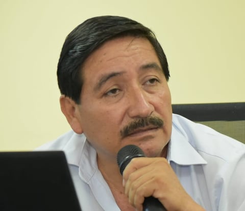 Man with dark hair and mustache wearing white shirt holding microphone, seated at desk