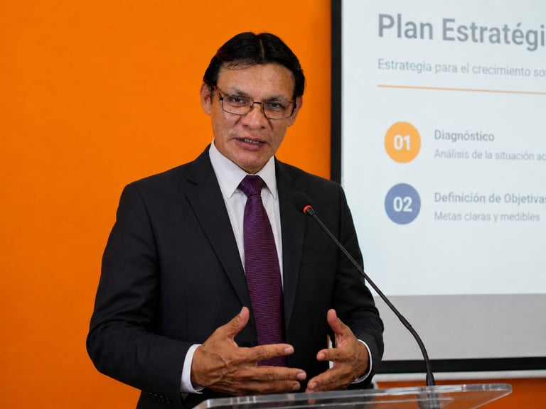 Man in business suit presenting at podium with orange and blue strategic plan slides visible behind him