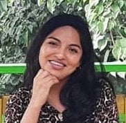 Woman with long black hair smiling at camera with hand near chin, ivy-covered wall background