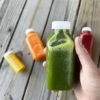 Hand holding a green juice bottle surrounded by colorful fresh juices on a wooden surface