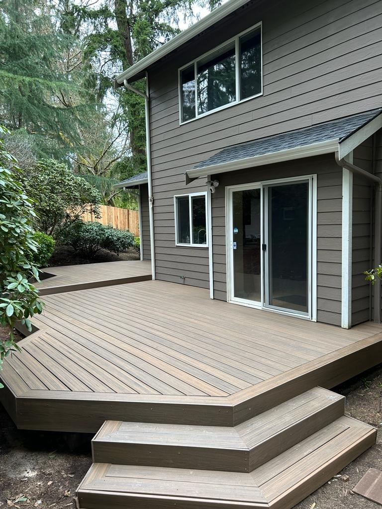 Modern gray house with composite deck, glass sliding doors, and wooden steps surrounded by trees and landscaping