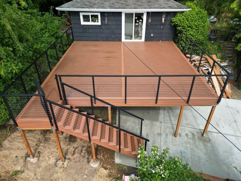New two-tier walnut composite deck with black cable railing attached to dark blue home, surrounded by green landscaping